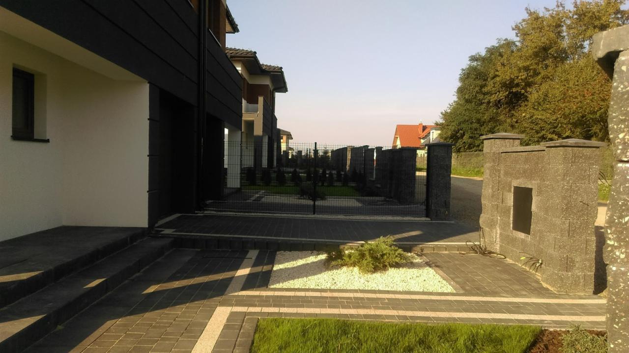 Nowoczesna posesja z czarnym ogrodzeniem panelowym i kamienną bramą, widok na zadbany trawnik, białe kamyki dekoracyjne i niskie iglaki wzdłuż ogrodzenia.