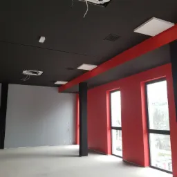 Wnętrze biura po remoncie, ściany pomalowane na czerwono i szaro, sufit czarny z panelami oświetleniowymi, widoczne okna i filary.