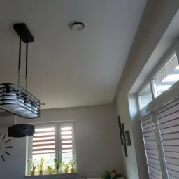 Gładka, biała powierzchnia sufitu w nowoczesnym wnętrzu, z widocznym okrągłym nawiewnikiem wentylacyjnym oraz designerską lampą wiszącą w stylu industrialnym.