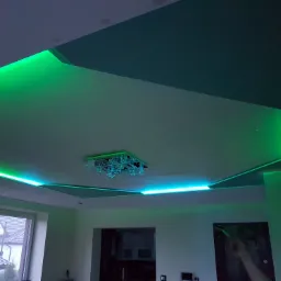 Nowoczesny sufit podwieszany z oświetleniem LED w kolorze zielonym, centralnie umieszczony żyrandol, widok z dołu na wykończone wnętrze.