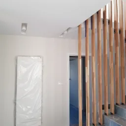 Nowoczesne wnętrze w trakcie wykańczania: białe ściany, drewniana ażurowa balustrada schodowa, marmurowe schody, drzwi zabezpieczone folią, minimalistyczne oświetlenie sufitowe.