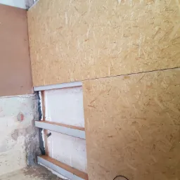 Montaż płyty OSB na ścianie z widoczną konstrukcją szkieletową wypełnioną pianką montażową, fragment starej ściany w tle.