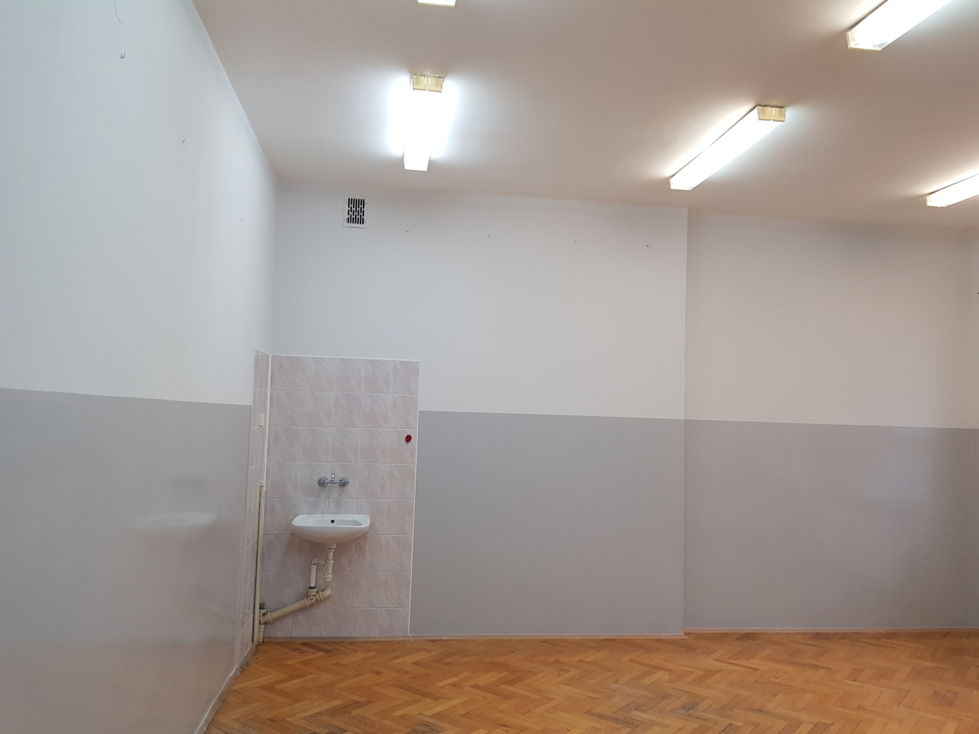 Wnętrze odnowionego pomieszczenia z pomalowanymi ścianami na dwa kolory - szary na dole i biały na górze, z widoczną umywalką z kafelkami w rogu i parkietem na podłodze.