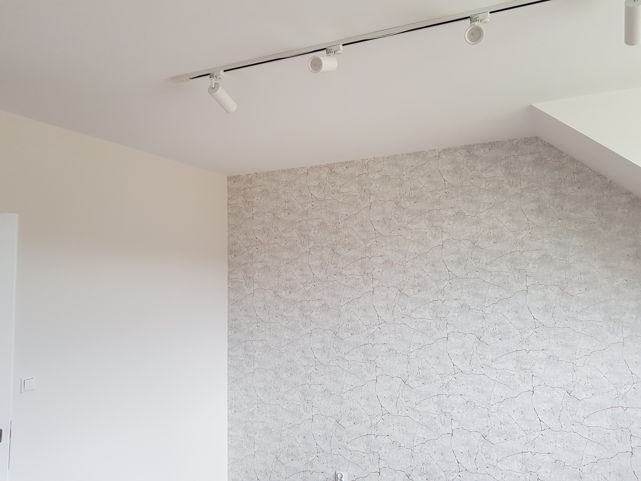 Wnętrze pokoju z tapetą imitującą spękaną powierzchnię, oświetlenie punktowe na szynie, białe ściany i sufit.