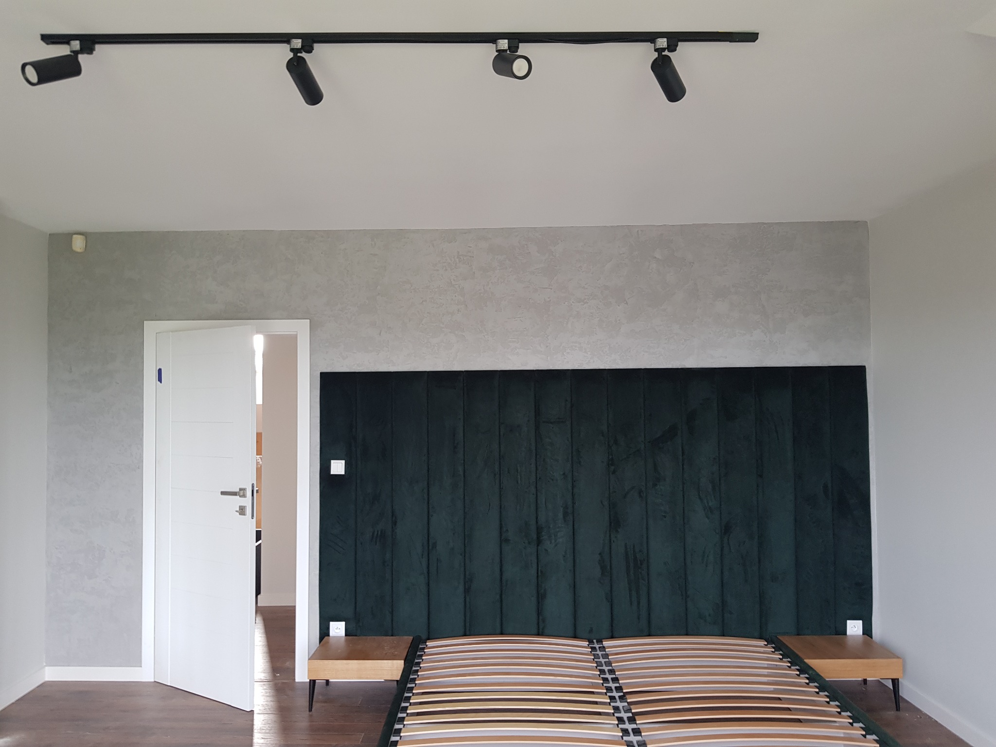 Wykończona sypialnia z dekoracyjną szarą ścianą imitującą beton, oświetleniem szynowym i zielonym panelem tapicerowanym za łóżkiem z drewnianym stelażem.