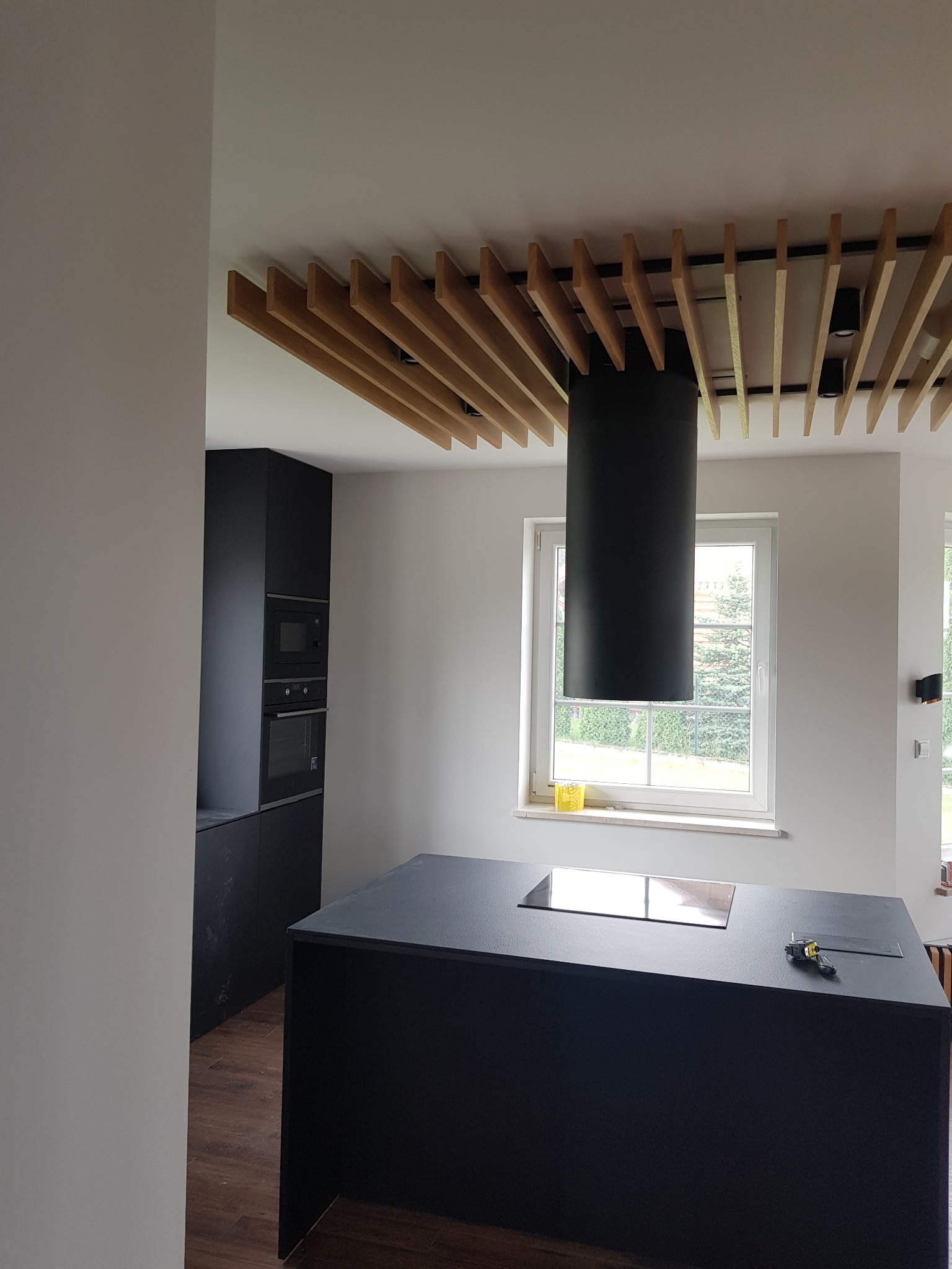 Nowoczesna kuchnia z czarną wyspą, zabudową i okapem cylindrycznym, oświetlona drewnianą konstrukcją sufitową.