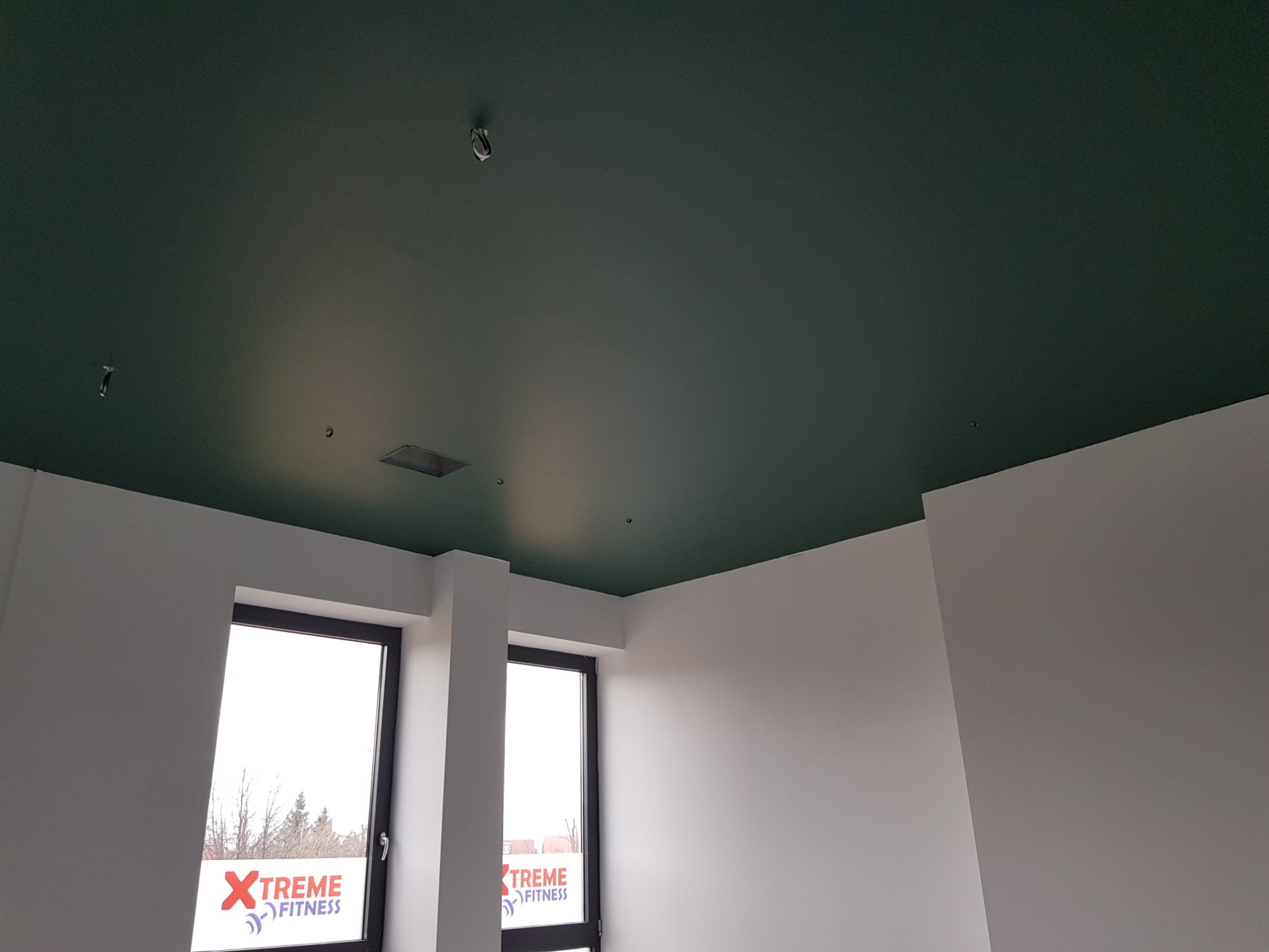 Pomieszczenie po remoncie z ciemnozielonym, świeżo pomalowanym sufitem i białymi ścianami, widoczne dwa okna z naklejkami reklamowymi Xtreme Fitness.