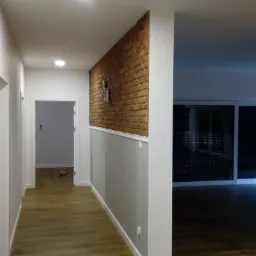 Korytarz po remoncie z panelami podłogowymi, białymi drzwiami i ścianą z cegły dekoracyjnej do połowy wysokości, poniżej pomalowana na szaro z białą listwą.