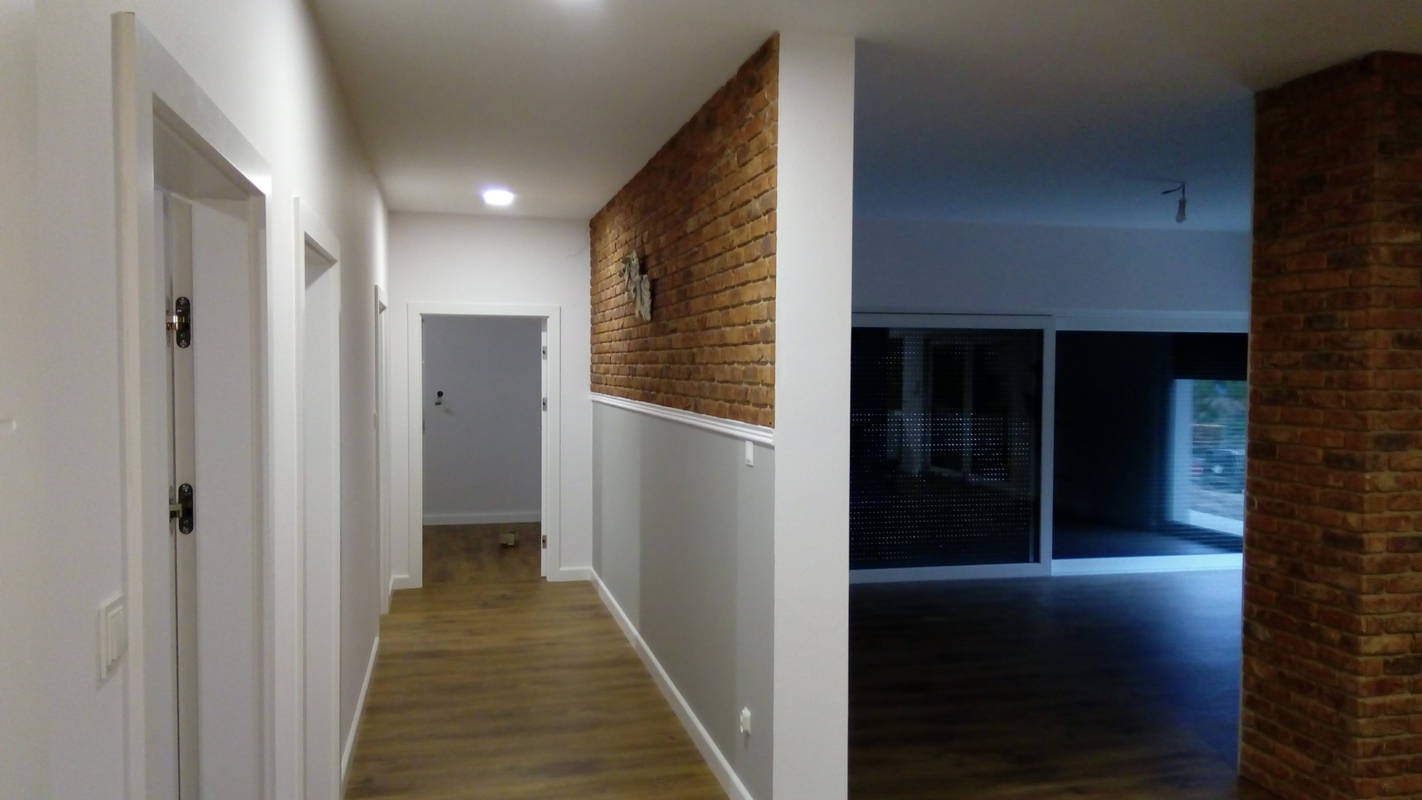 Korytarz po remoncie z panelami podłogowymi, białymi drzwiami i ścianą z cegły dekoracyjnej do połowy wysokości, poniżej pomalowana na szaro z białą listwą.