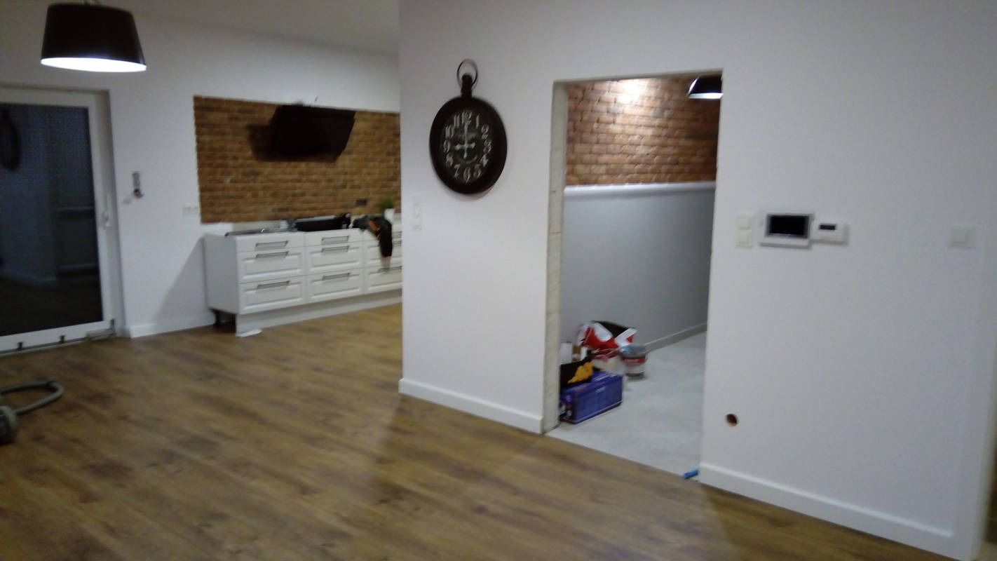 Widok na fragment wyremontowanego mieszkania z drewnianą podłogą, białą zabudową kuchenną z ceglaną ścianą w tle, ozdobnym zegarem na ścianie i widocznymi śladami po remoncie w przejściu do innego...