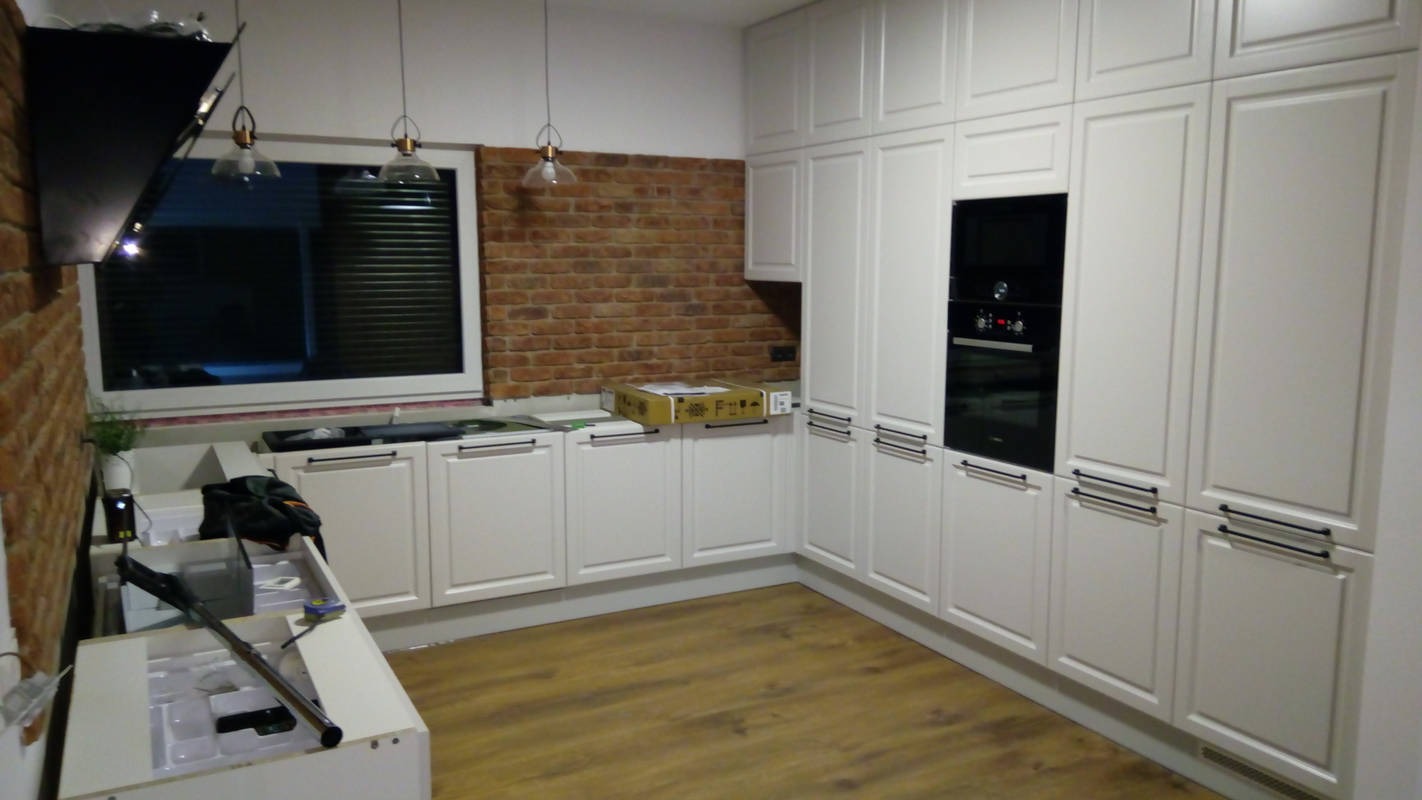 Wnętrze kuchni w trakcie montażu: białe szafki z czarnymi uchwytami, ceglana ściana, okno z żaluzjami, lampy wiszące i podłoga z paneli.