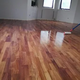 Lakierowana podłoga z drewna egzotycznego z widocznym wałkiem malarskim podczas aplikacji lakieru, odbicie okna w mokrej powierzchni.