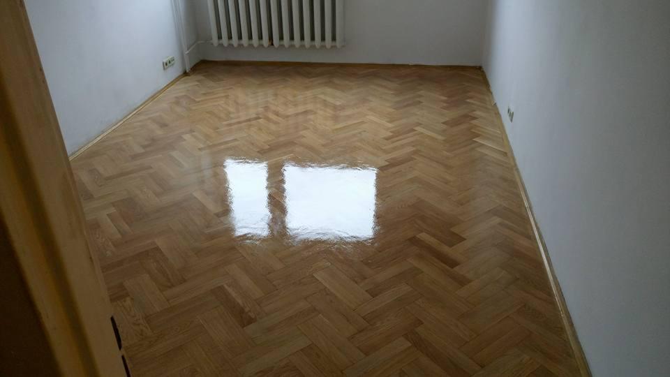 Świeżo polakierowany parkiet ułożony w jodełkę, z wyraźnym odbiciem okna, w pokoju z białymi ścianami i grzejnikiem żeberkowym.