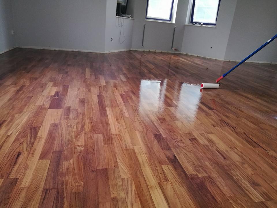 Lakierowana podłoga z drewna egzotycznego z widocznym wałkiem malarskim podczas aplikacji lakieru, odbicie okna w mokrej powierzchni.