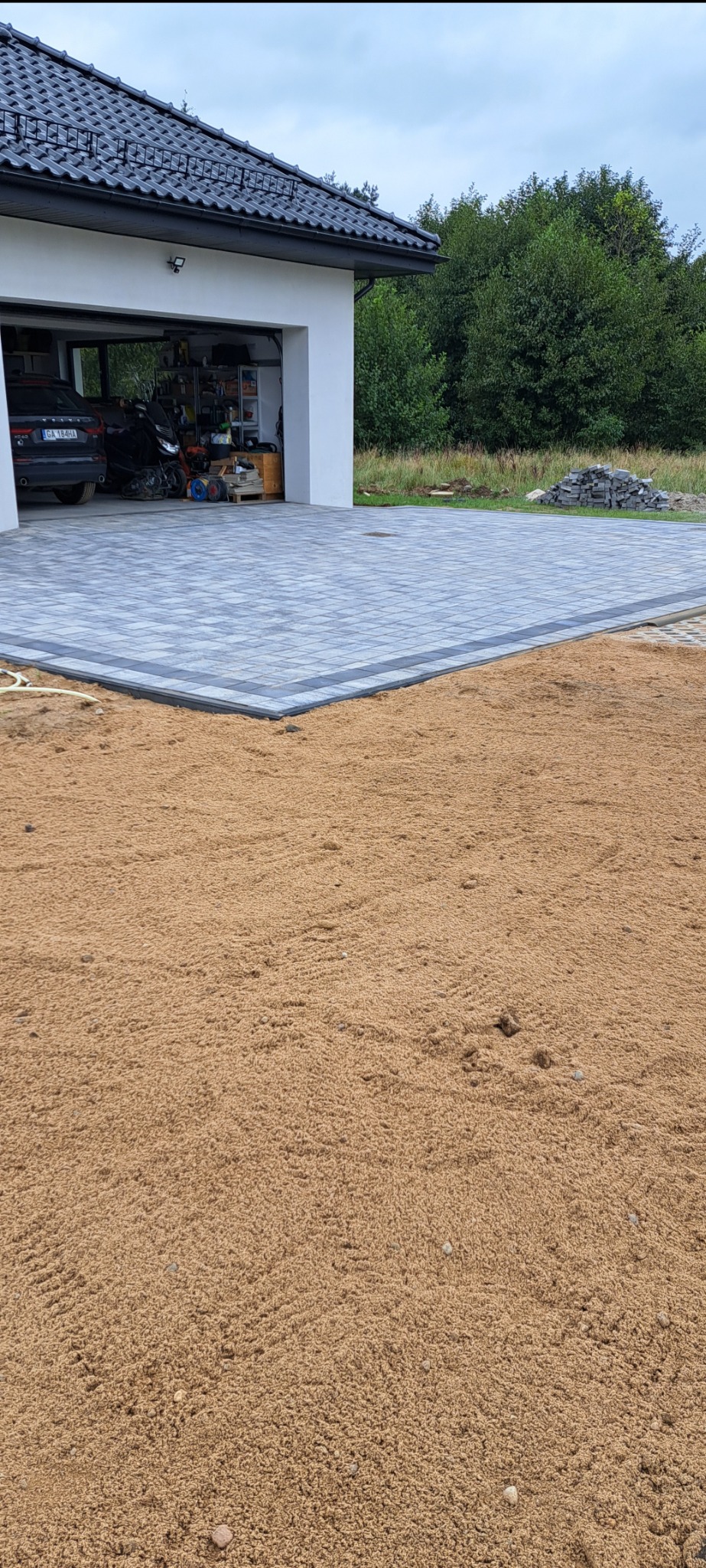 Układanie kostki brukowej w kolorze szarości z ciemniejszym obramowaniem, widoczny fragment podsypki piaskowej, garaż z samochodem w tle.