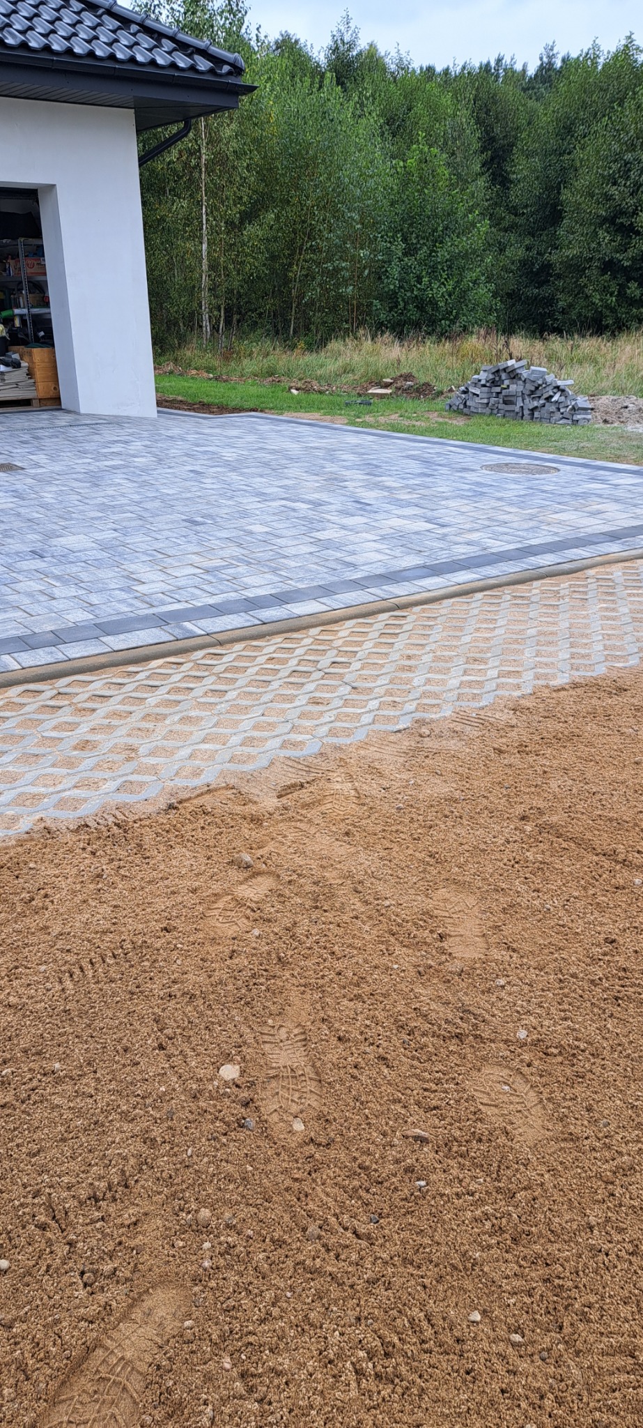 Częściowo ułożona, szara kostka brukowa na podjeździe, z widoczną warstwą piasku i śladami stóp, w tle dom z czarnym dachem i zielone drzewa.