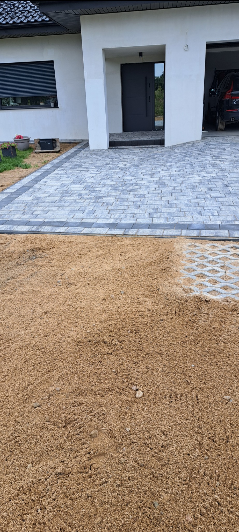 Częściowo ułożona szara kostka brukowa przed domem z szarą stolarką okienną i drzwiową, widoczny piasek i ażurowe płyty betonowe.