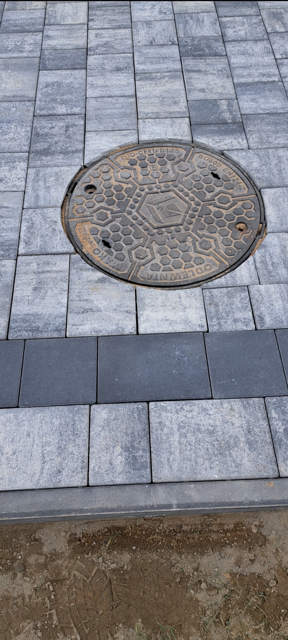 Szara kostka brukowa ułożona wokół okrągłej, żeliwnej studzienki z widocznym napisem 'MAKO ODLEWNIA'. W tle widoczna linia ciemniejszej kostki.