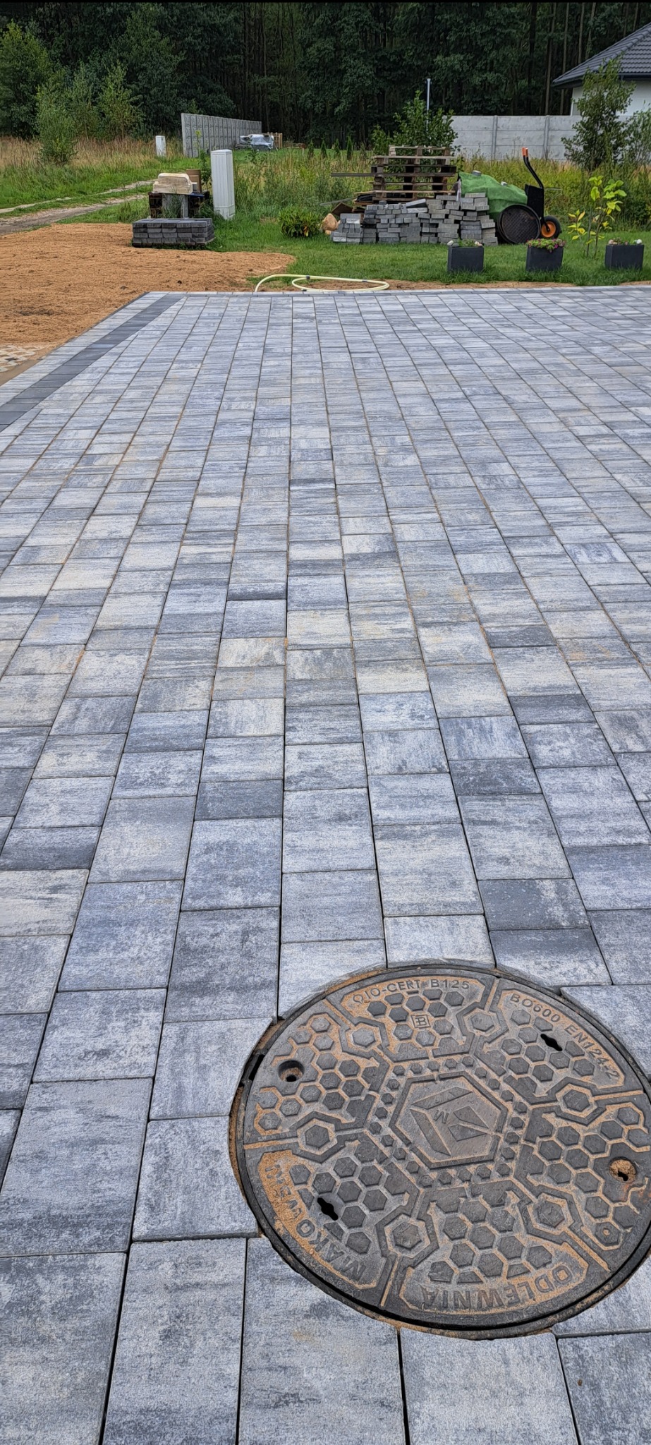 Ułożona szara kostka brukowa z widocznym włazem kanalizacyjnym w prawym dolnym rogu, w tle trawnik i drzewa.