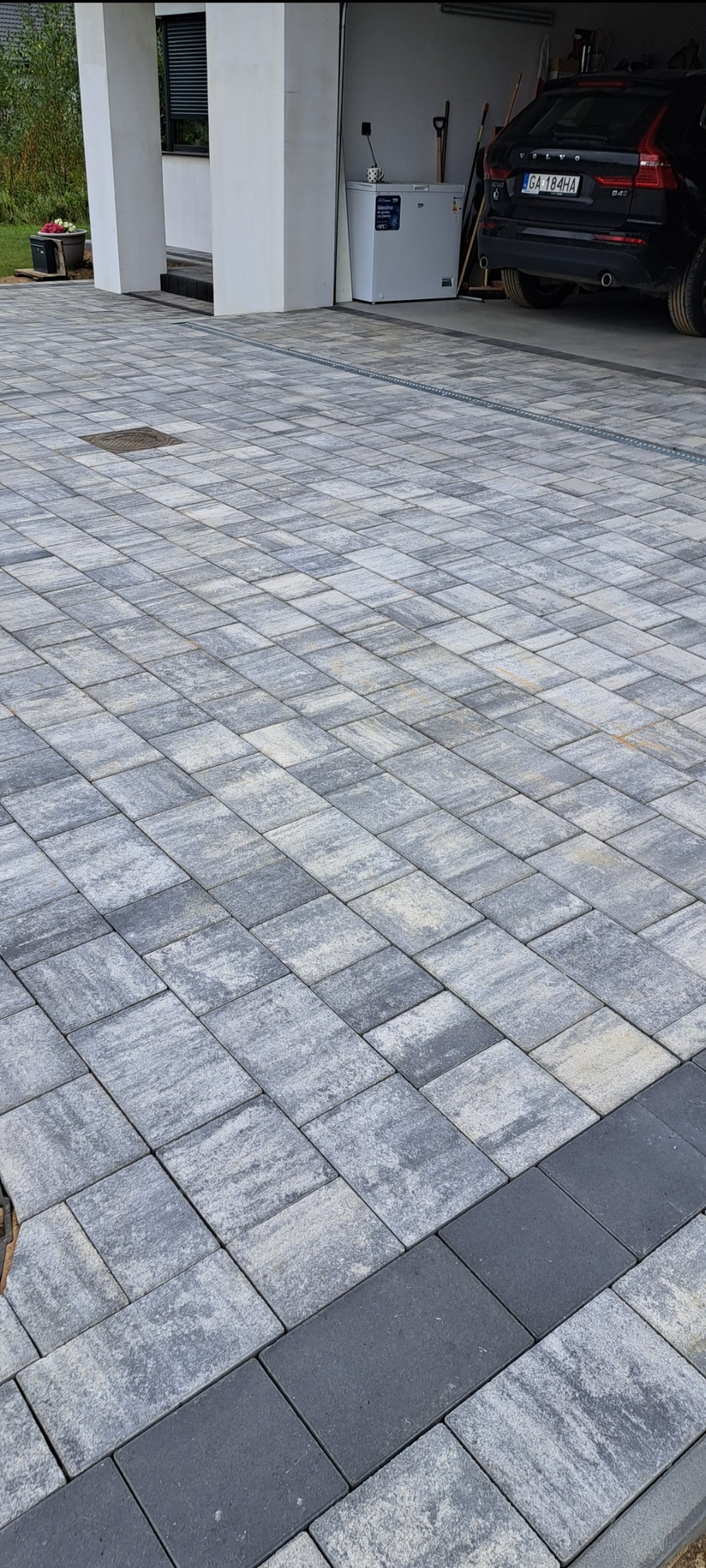 Nowoczesny podjazd z szarej kostki brukowej ułożonej we wzór prostokątów, z ciemnoszarym obramowaniem, widoczny fragment domu z garażem i samochodem w tle.