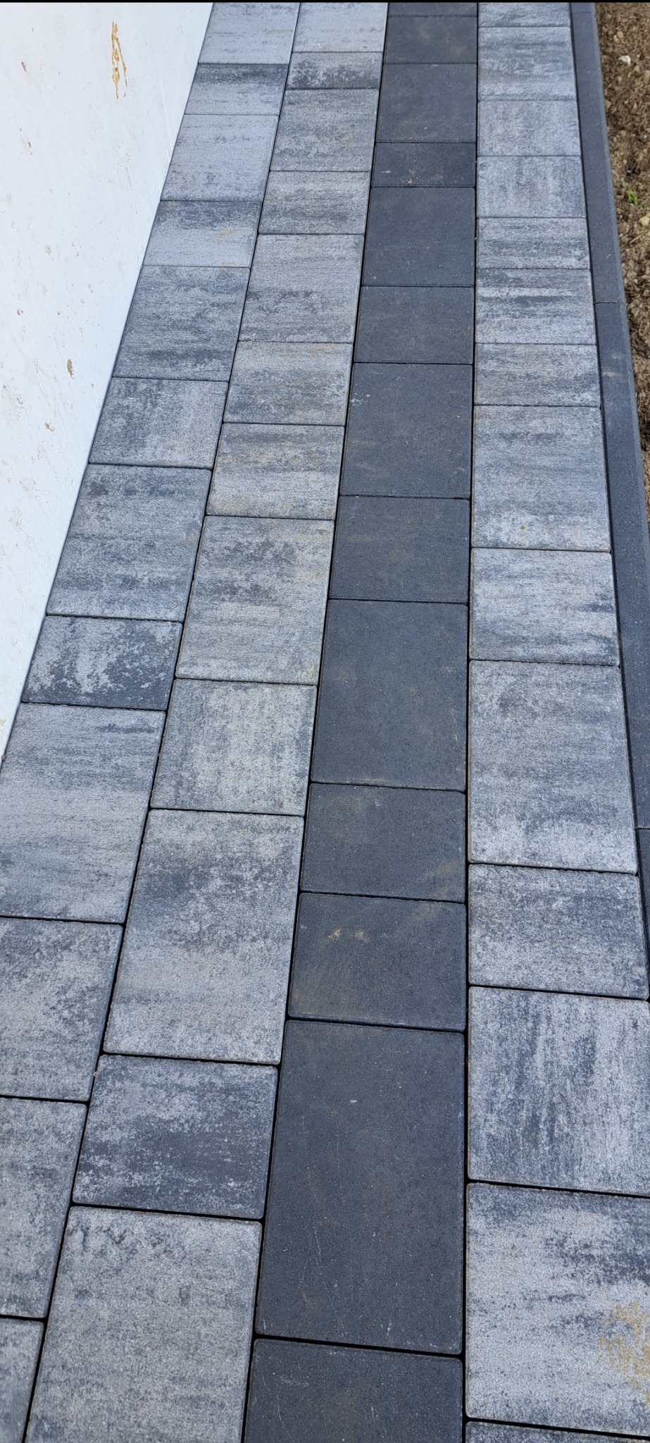 Ułożona ścieżka z szarej kostki brukowej z ciemnoszarym pasem pośrodku, przylegająca do białej ściany i ziemi.