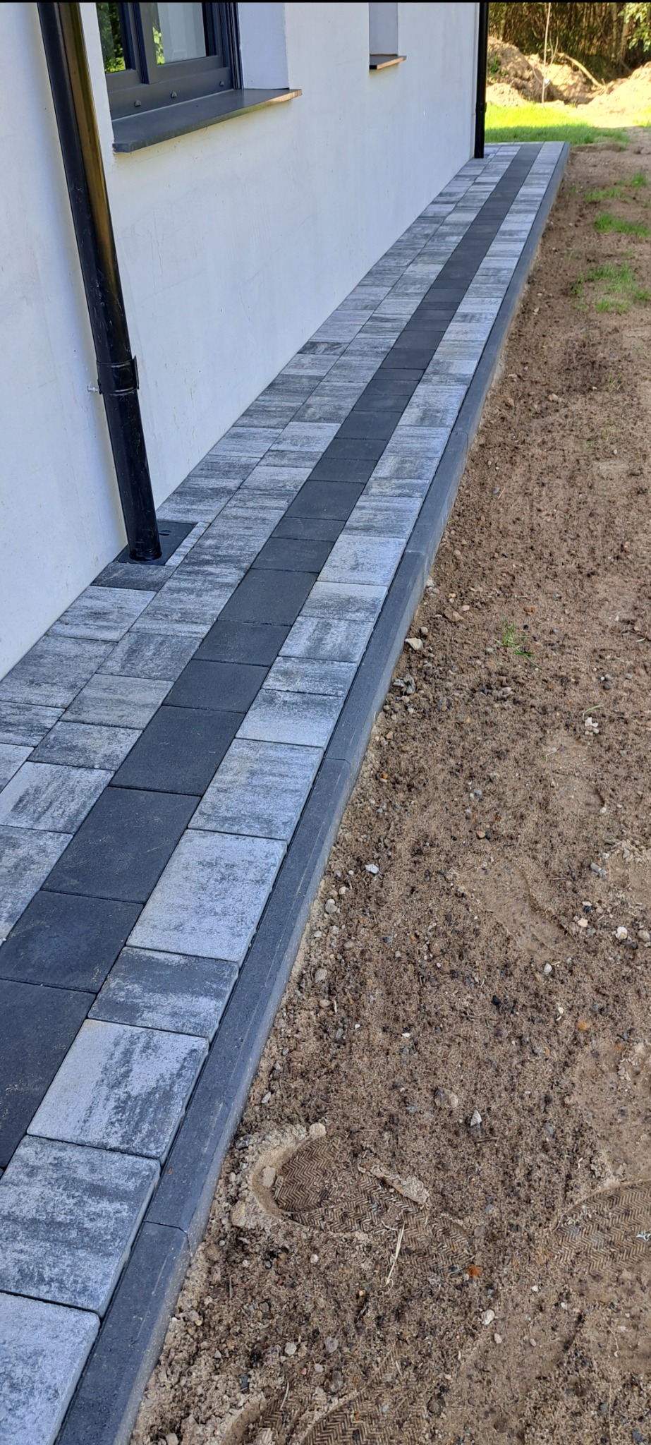 Ułożona ścieżka z kostki brukowej w dwóch odcieniach szarości biegnąca wzdłuż białej ściany budynku z widoczną rynną spustową i śladami butów w piasku obok.