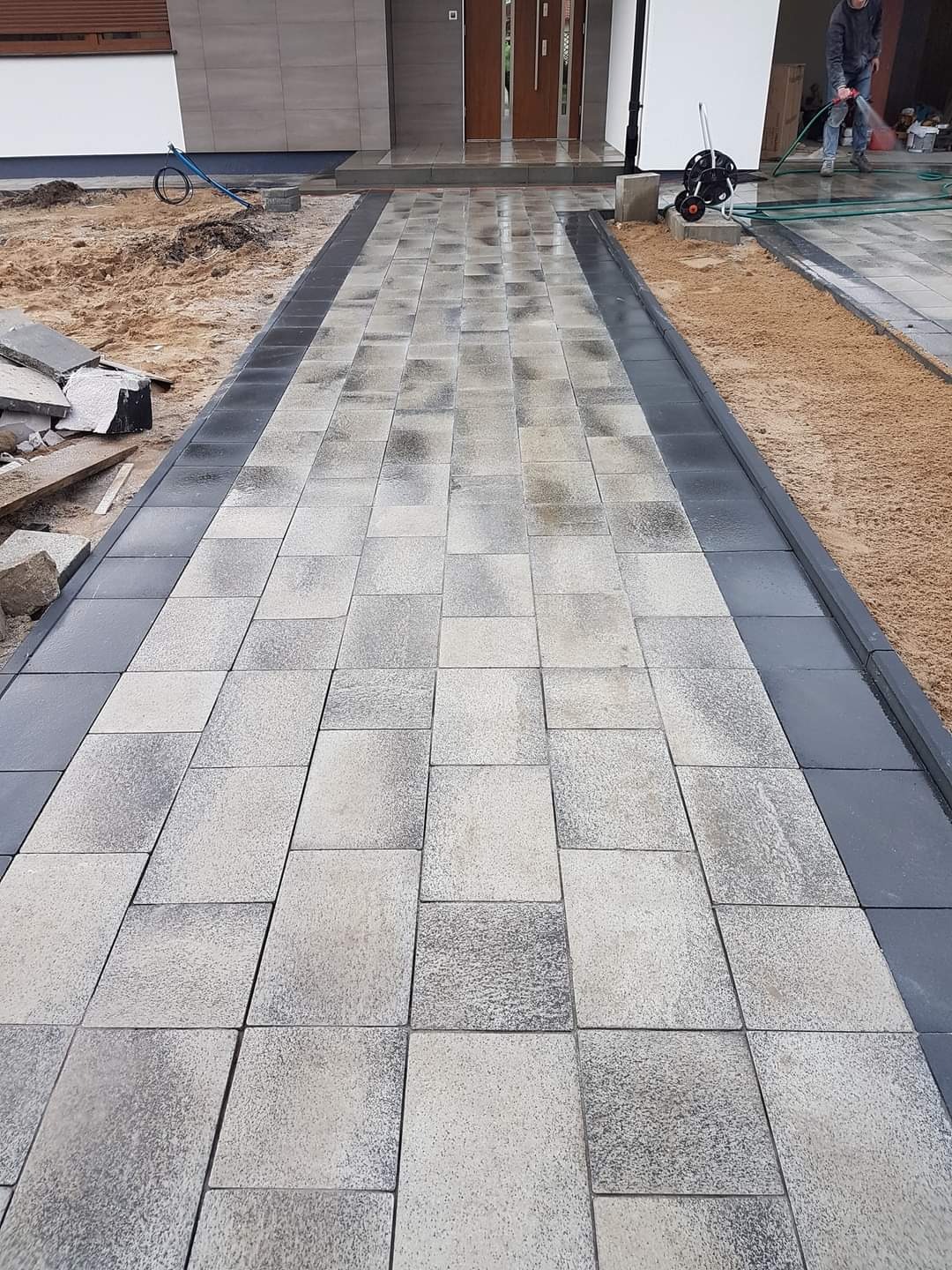Świeżo ułożona, wilgotna kostka brukowa prowadząca do domu, z ciemnoszarym obramowaniem, widoczny fragment elewacji, osoba spłukująca resztki piasku w tle.