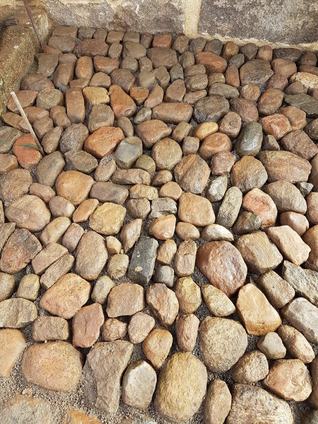 Wykończona powierzchnia z nieregularnych, brązowo-szarych kamieni rzecznych, ułożonych w zaprawie piaskowej przy ścianie budynku.