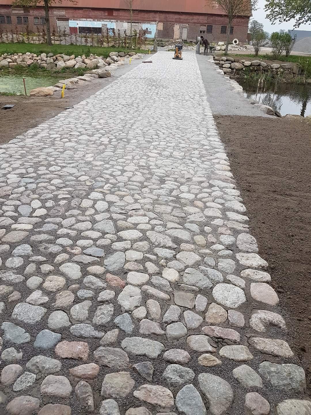 Ścieżka z nieregularnej kostki granitowej w ogrodzie, obok stawu, z ubijarką w tle.