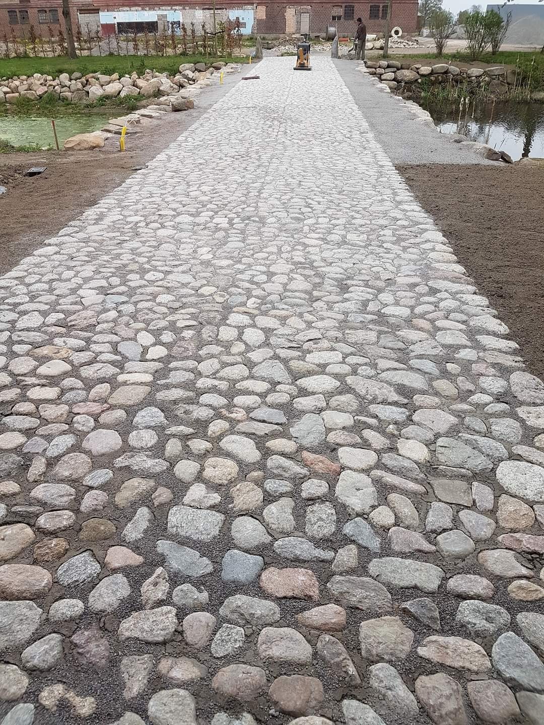 Długa ścieżka z nieregularnych, jasnych kamieni polnych, w tle budynek i staw, widoczna ubijarka w oddali.