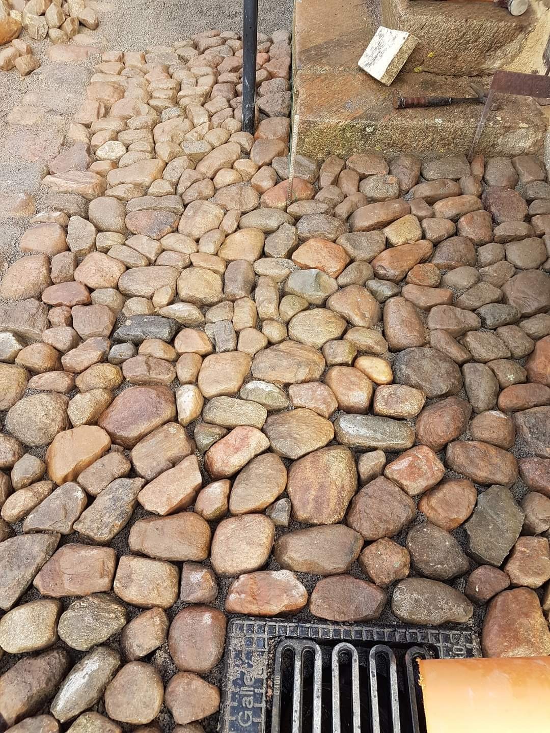 Ścieżka z zaokrąglonych kamieni w odcieniach brązu i szarości, prowadząca do schodów z kamienia, z widoczną kratką odpływową w dolnej części zdjęcia.