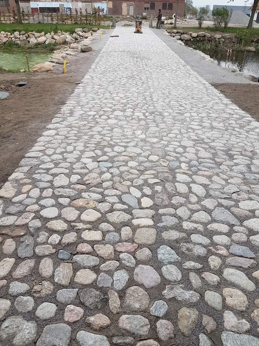 Długa ścieżka wyłożona nieregularnymi kamieniami granitowymi, prowadząca przez park z elementami wodnymi, w tle budynek i pracownik z ubijarką.