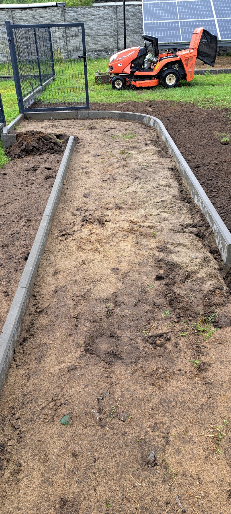 Przygotowanie podłoża pod ścieżkę z obrzeżami z szarej kostki, widoczna pomarańczowa kosiarka traktorek na trawniku w tle.