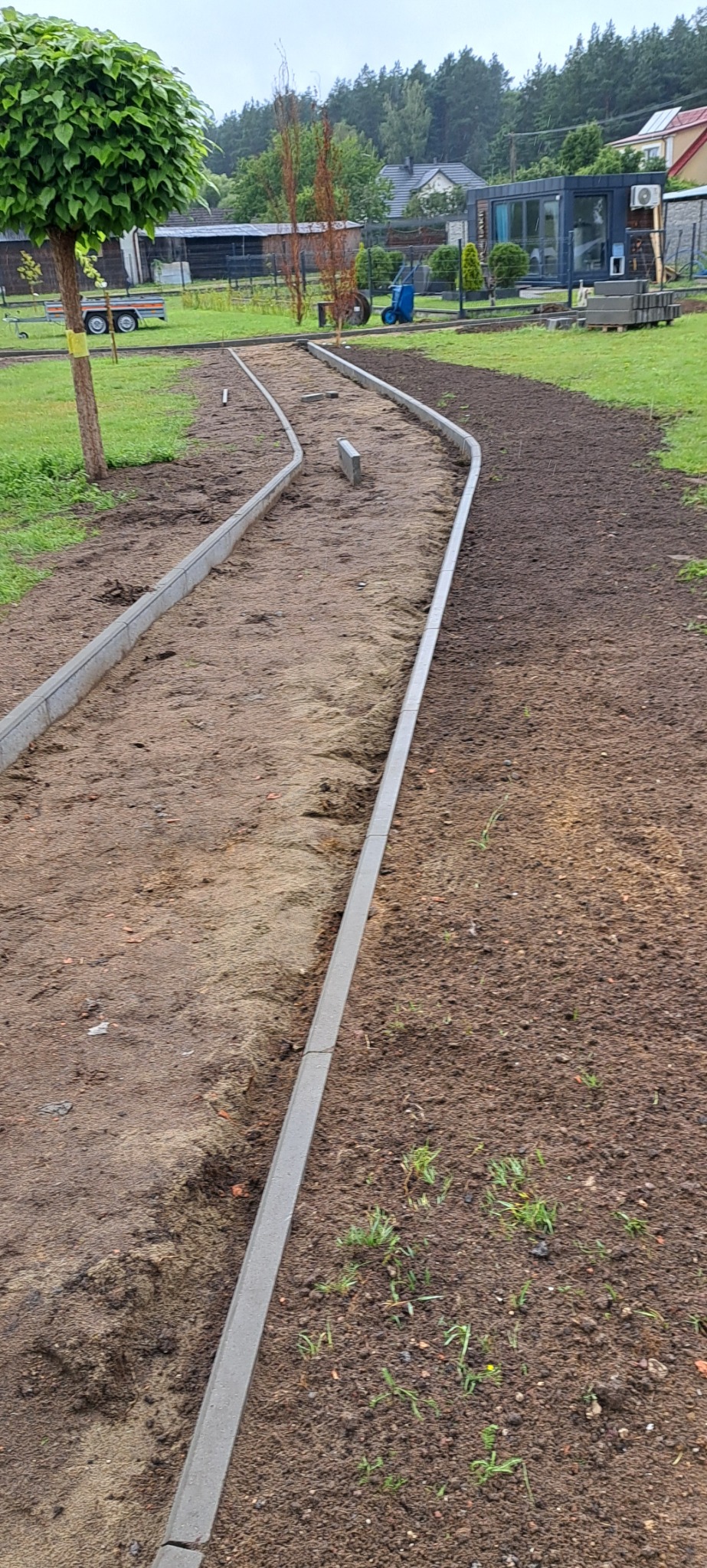 Przygotowanie podłoża pod ścieżkę z obrzeżami z szarej kostki betonowej, widoczne podłoże z piasku i ziemi, w tle drzewa i zabudowania.