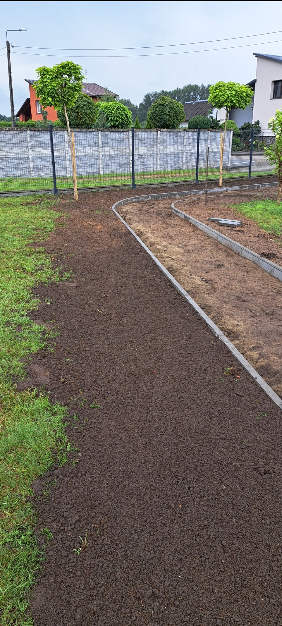 Przygotowany teren pod ścieżkę z obrzeżami z kostki betonowej, widoczne wyrównane podłoże i fragmenty trawnika, w tle ogrodzenie panelowe i drzewa.