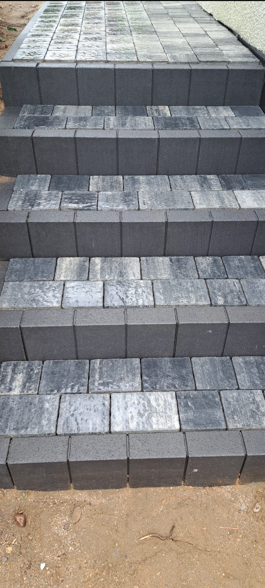 Schody zewnętrzne wyłożone szarą kostką brukową o zróżnicowanej fakturze, z widocznymi fugami i krawężnikami, na tle wilgotnej ziemi.
