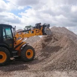 Żółto-czarna koparko-ładowarka JCB nabiera żwir z dużej pryzmy; w tle widoczny dom z czerwonym dachem i pochmurne niebo.