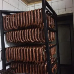 Rzędy wędzonych kiełbas zwisające z metalowego regału w chłodni, widoczne białe kafelki na ścianach i ciemne pomieszczenie w tle.