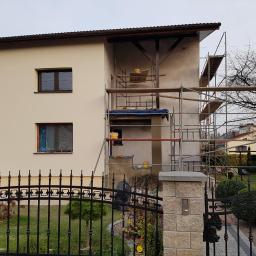 RomanDepta - Dwukondygnacyjny dom w trakcie prac elewacyjnych: część ściany pomalowana na kremowo, druga część w szarym tynku, rusztowania ustawione przy ścianie, okno zabezpieczone folią, widoczna brama...