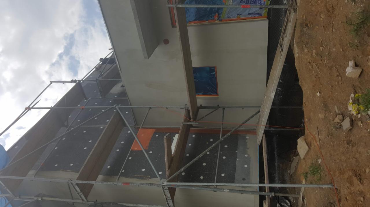 Dom w trakcie ocieplania, widoczne rusztowania, płyty izolacyjne z białymi kołkami i fragment świeżo otynkowanej ściany z zabezpieczonymi oknami.