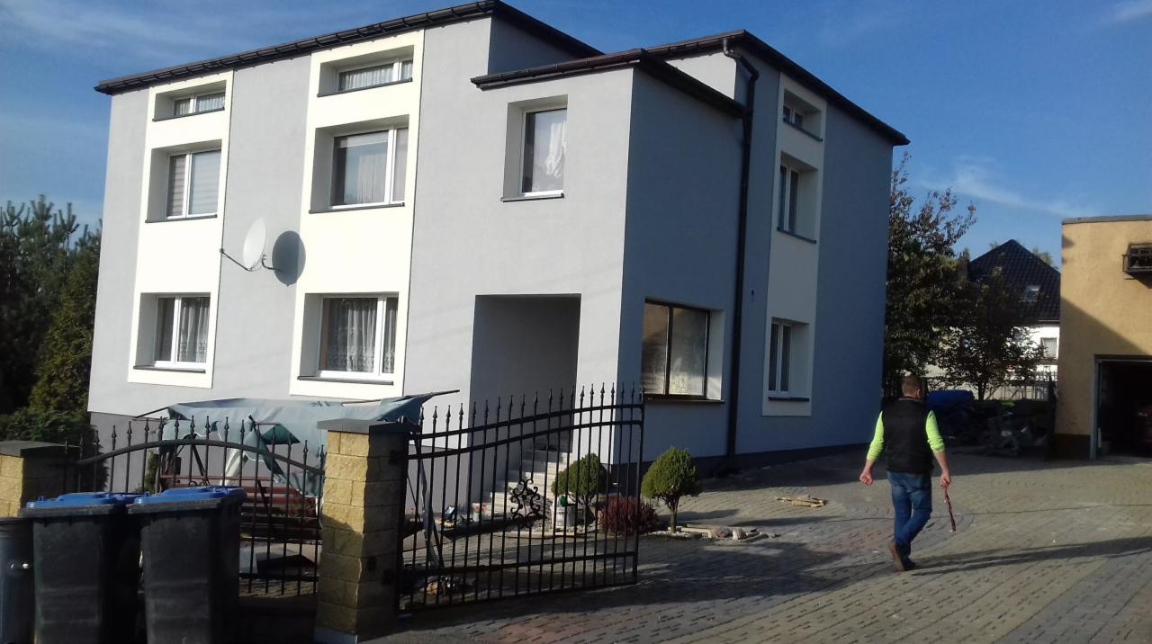 Dwupiętrowy dom po remoncie elewacji, pomalowany na szaro z białymi akcentami wokół okien, widoczny z zewnątrz przez czarne metalowe ogrodzenie i kostkę brukową, z satelitą na ścianie i mężczyzną...