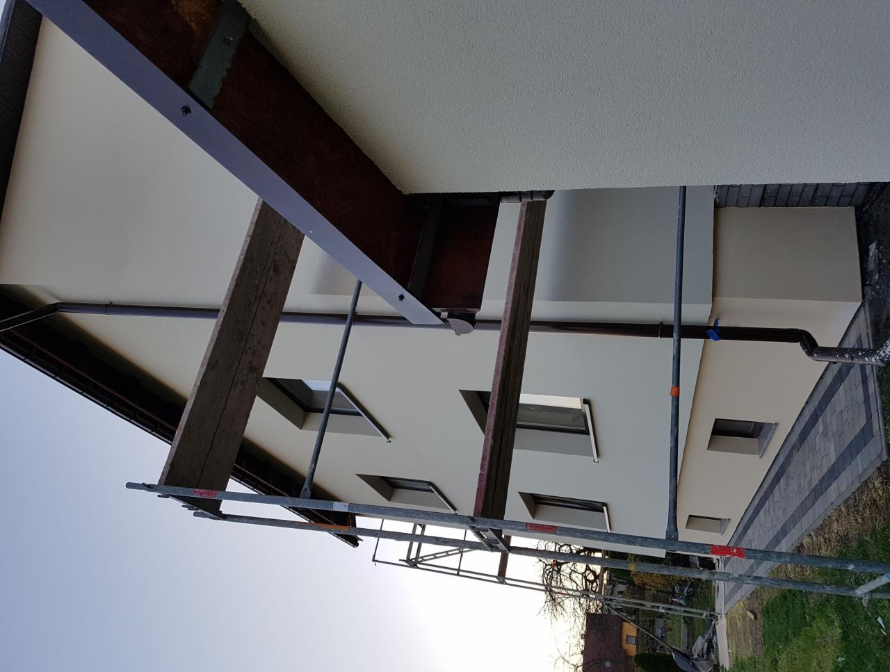 Dom jednorodzinny w trakcie prac elewacyjnych, widoczne rusztowania, świeżo otynkowana ściana w odcieniach beżu, fragment dachu z drewnianymi elementami konstrukcyjnymi.