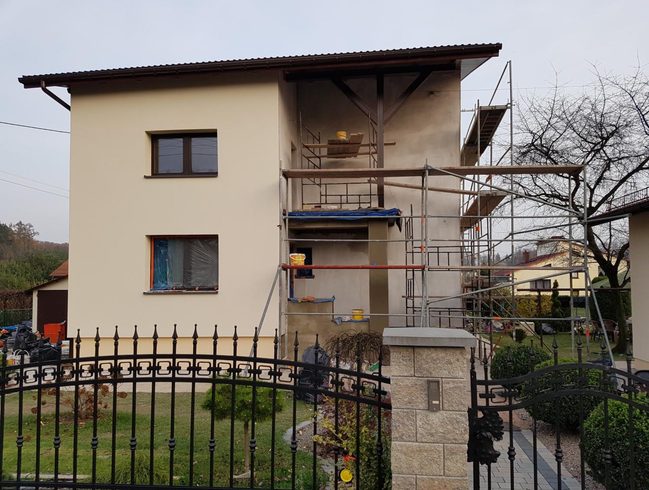 Dwukondygnacyjny dom w trakcie prac elewacyjnych: część ściany pomalowana na kremowo, druga część w szarym tynku, rusztowania ustawione przy ścianie, okno zabezpieczone folią, widoczna brama...