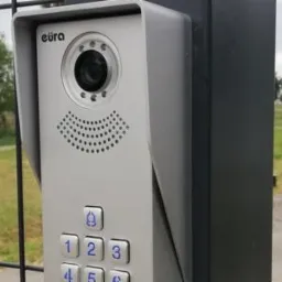Szczegółowe ujęcie wideodomofonu Eura z klawiaturą numeryczną, zamontowanego na czarnym słupku bramy, z widokiem na zielone pole w tle.