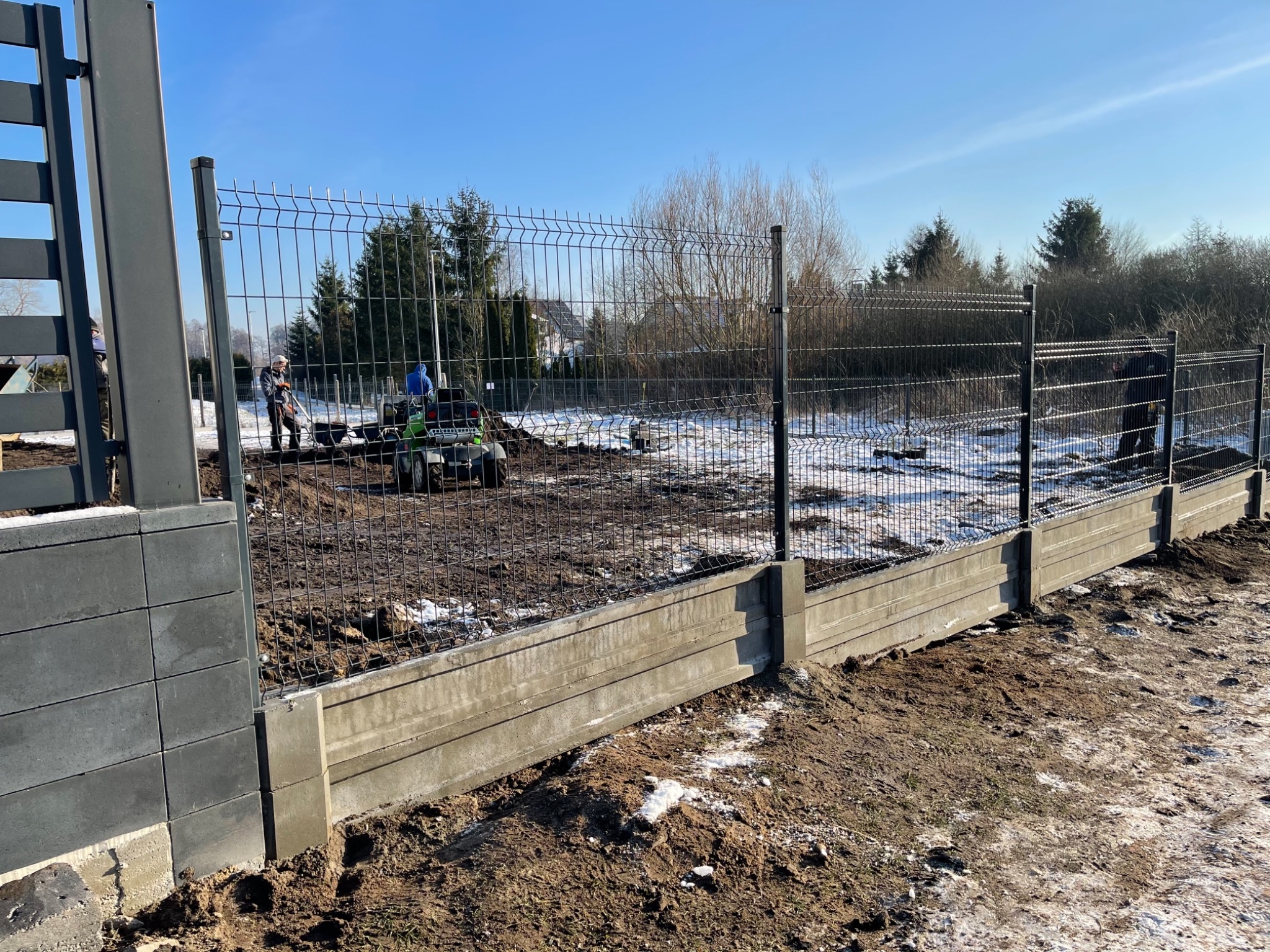 Montaż panelowego ogrodzenia na betonowej podmurówce; widoczni pracownicy z taczką i mini-ładowarką na tle zaśnieżonego ogrodu i drzew.