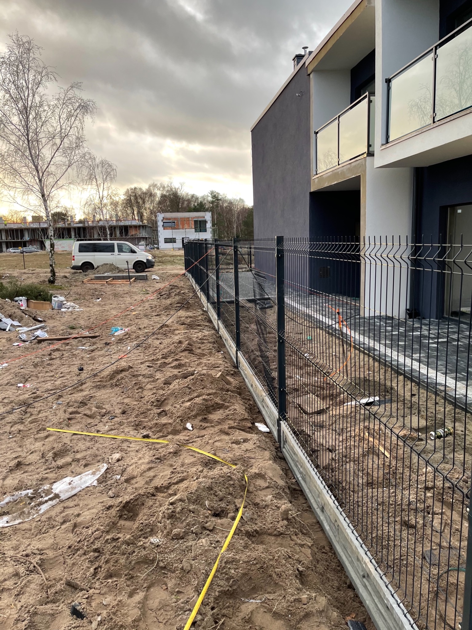 Metalowe ogrodzenie panelowe z podmurówką betonową w trakcie montażu, widoczne rozłożone narzędzia i taśma miernicza na piaszczystym terenie budowy, w tle budynek mieszkalny w nowoczesnym stylu.