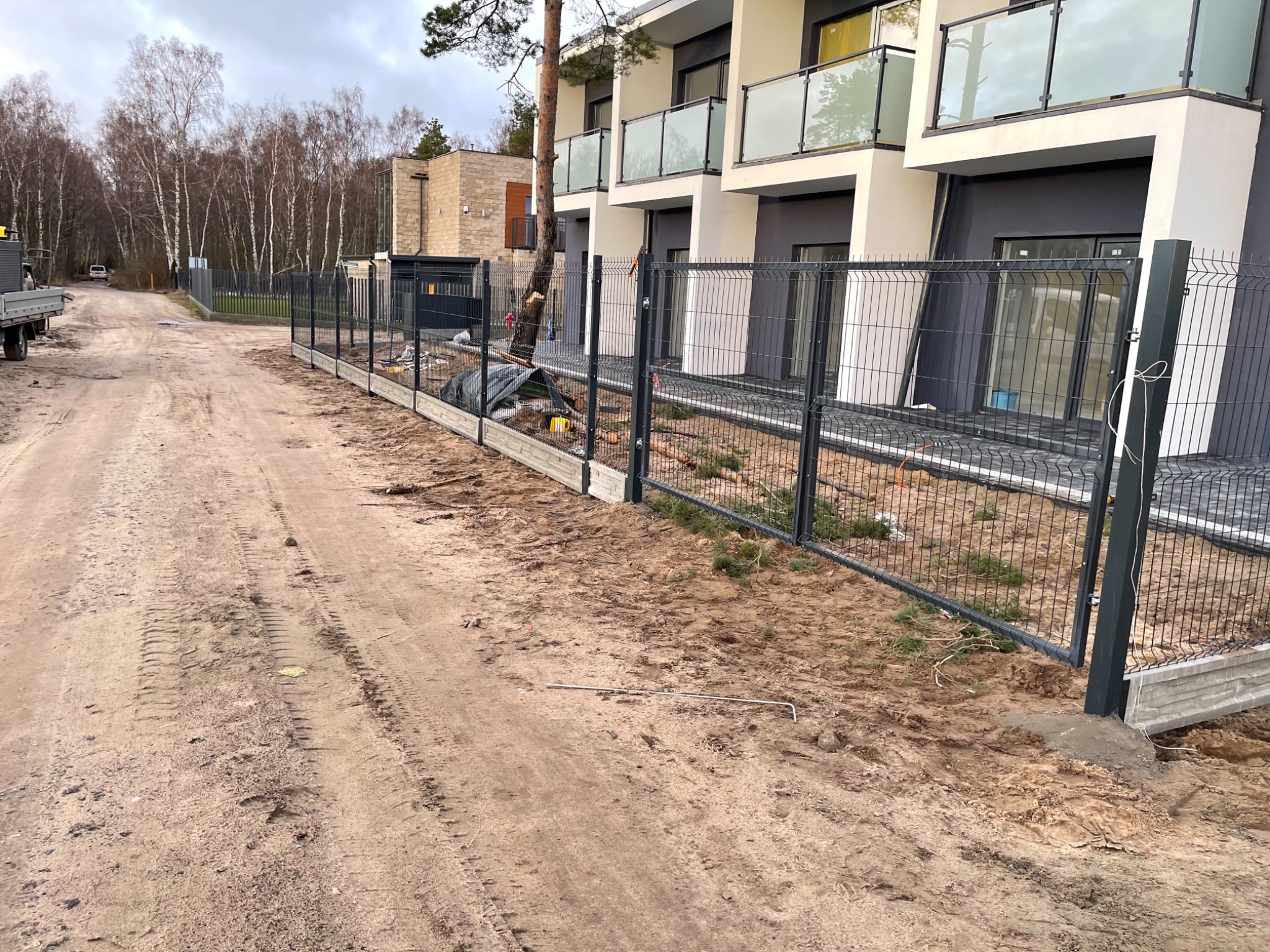 Montaż panelowego ogrodzenia z podmurówką betonową przy nowoczesnym budynku wielorodzinnym, widoczne ślady prac ziemnych i narzędzie na piaszczystej drodze.