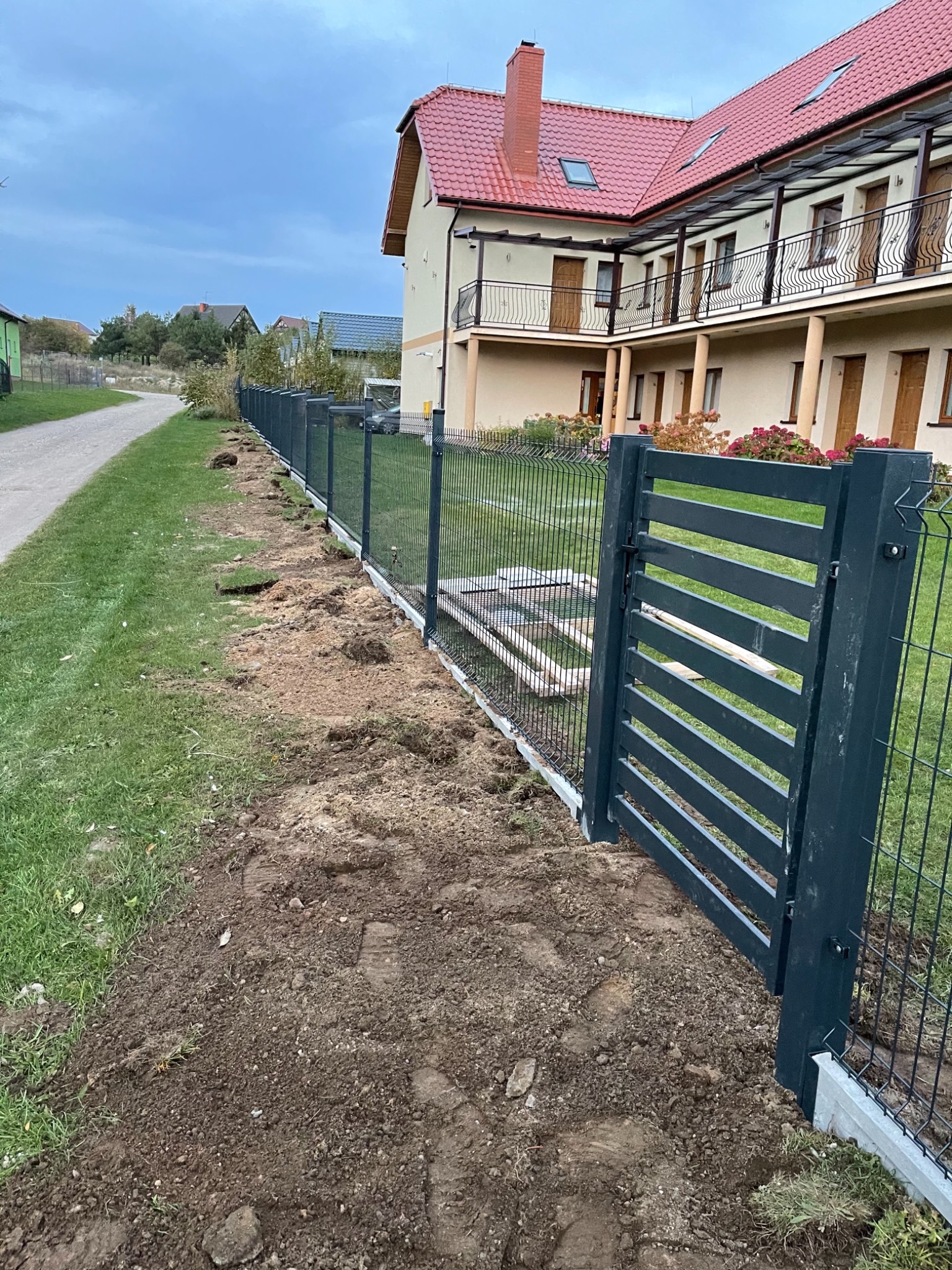 Nowoczesne, grafitowe ogrodzenie panelowe z poziomą bramą wjazdową, widoczne ślady prac ziemnych po montażu, w tle dwupiętrowy dom z czerwonym dachem.