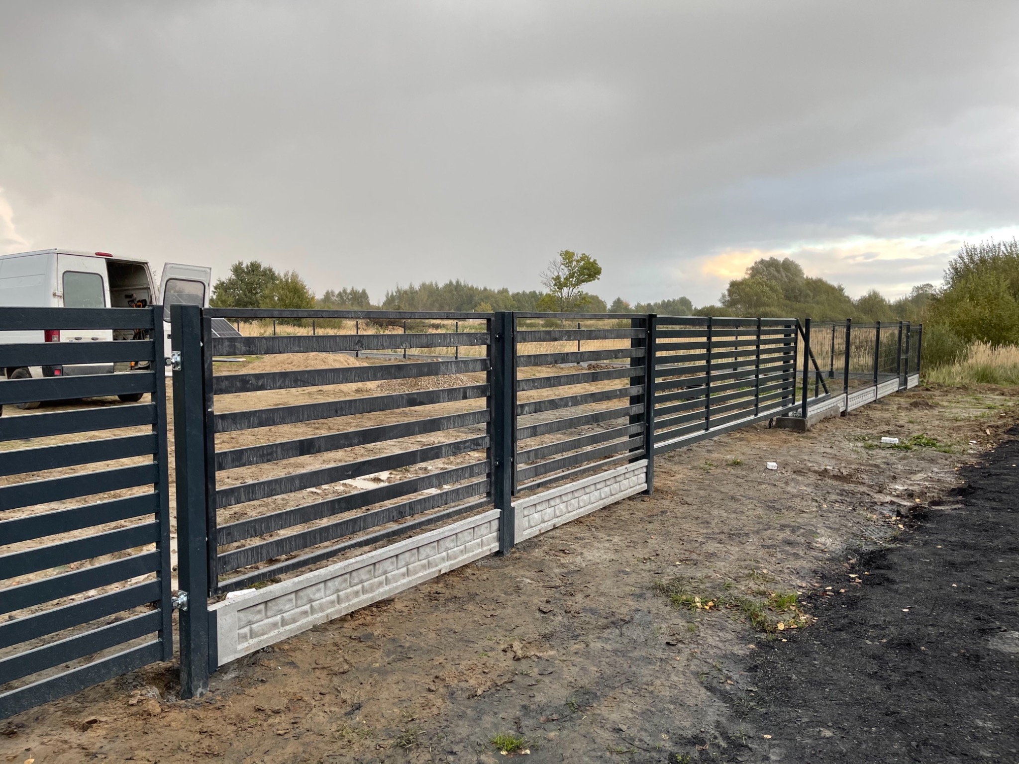 Nowoczesne, poziome ogrodzenie panelowe w kolorze antracytowym z betonową podmurówką, widok z zewnątrz posesji, z otwartym białym busem w tle.