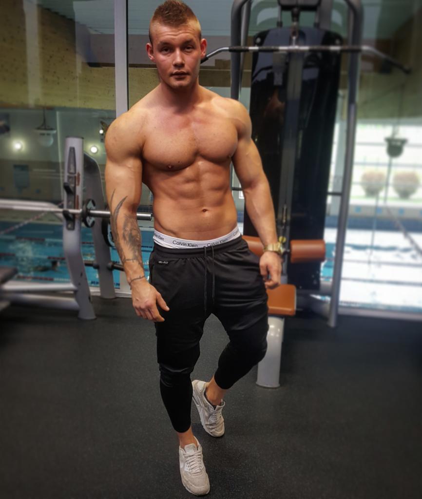 Mężczyzna o muskularnej sylwetce pozuje w siłowni, widoczny tatuaż na ramieniu, w tle sprzęt do ćwiczeń i basen.